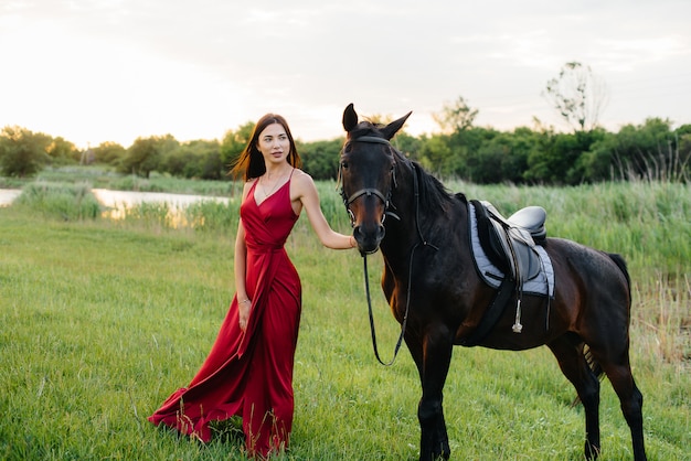 Une jeune jolie fille dans une robe rouge pose sur un ranch avec un étalon pur-sang au coucher du soleil. Aime et soigne les animaux.
