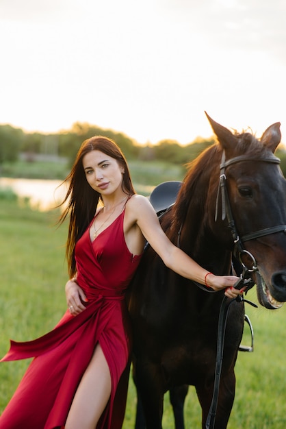 Une jeune jolie fille dans une robe rouge pose sur un ranch avec un étalon pur-sang au coucher du soleil. Aime et soigne les animaux.