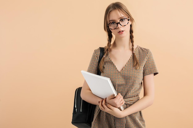 Jeune jolie fille avec deux tresses en combinaison de tweed et lunettes avec sac à dos noir sur l'épaule tenant des blocs-notes dans les mains tout en regardant pensivement à huis clos sur fond beige