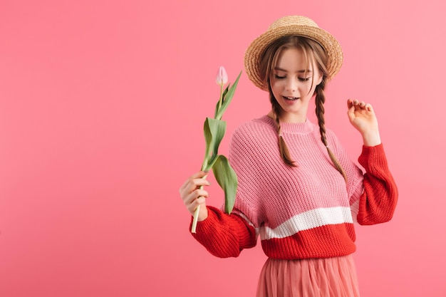 Jeune jolie fille avec deux tresses en pull et chapeau de paille tenant une tulipe à la main tout en dansant joyeusement sur fond rose