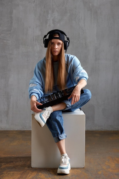 Une jeune jolie fille dj aux cheveux longs dans un jean pull bleu et une casquette de baseball noire pose tout en s'asseyant