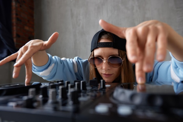 Une jeune jolie fille dj aux cheveux longs dans un pull bleu et une casquette de baseball noire pose avec un mix dj noir