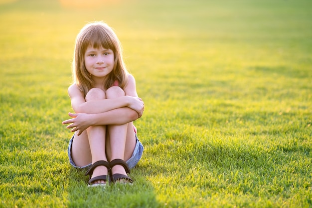 Jeune jolie fille enfant assise sur une pelouse d'herbe verte fraîche par une chaude journée d'été à l'extérieur.