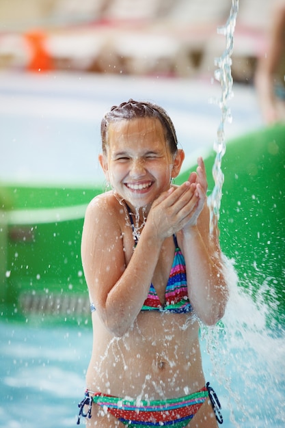 Jeune Jolie Fille En Maillot De Bain Rayé Coloré Rit, Se Tient Sous Une Cascade Dans Un Parc Aquatique.