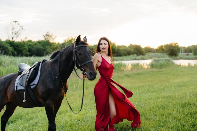 Une jeune jolie fille en robe rouge pose sur un ranch avec un étalon pur-sang au coucher du soleil. Aimer et prendre soin des animaux.