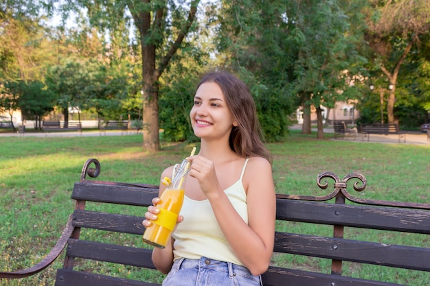 Jeune jolie fille se repose sur le banc dans le parc et boit du jus de fruits