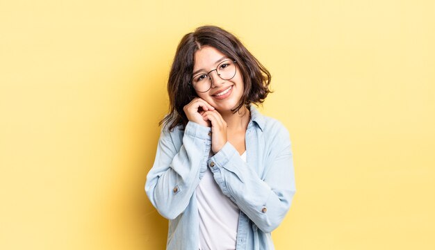 Jeune jolie fille se sentant amoureuse et ayant l'air mignonne, adorable et heureuse, souriant de manière romantique avec les mains à côté du visage