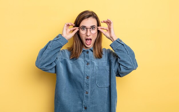 Jeune jolie fille se sentant choquée, étonnée et surprise, tenant des lunettes avec un regard étonné et incrédule