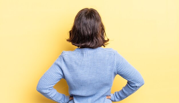 Jeune jolie fille se sentant confuse ou pleine ou des doutes et des questions, se demandant, avec les mains sur les hanches, vue arrière