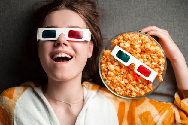 Jeune jolie fille se trouve sur un fond gris dans des lunettes 3d mange du popcrone et regarde un film