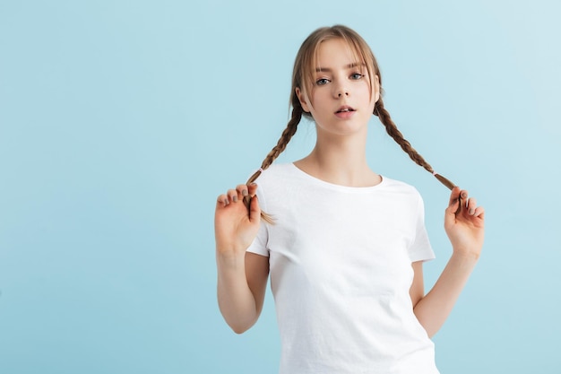 Jeune jolie fille en t-shirt blanc tenant deux tresses aux cheveux dans les mains tout en regardant rêveusement à huis clos sur fond bleu