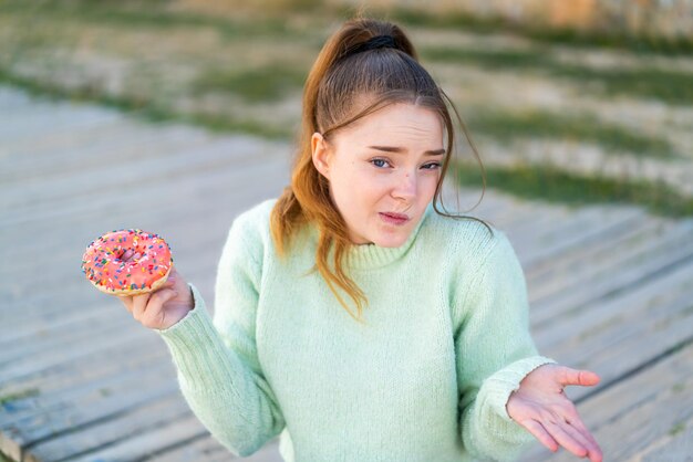 Jeune jolie fille tenant un beignet à l'extérieur faisant un geste de doute tout en levant les épaules