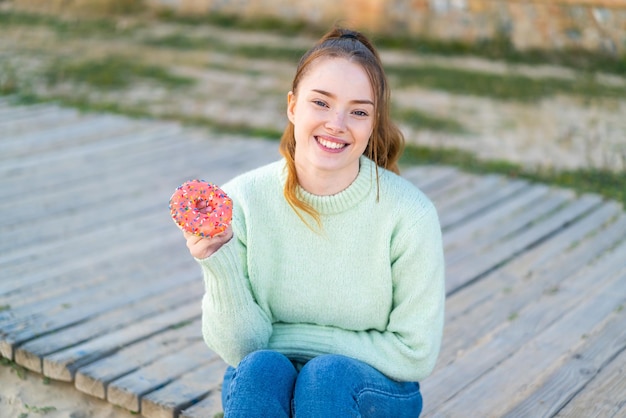 Jeune jolie fille tenant un beignet à l'extérieur souriant beaucoup