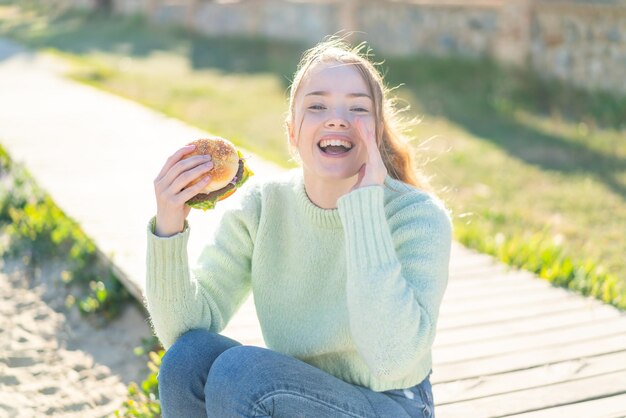 Jeune jolie fille tenant un hamburger à l'extérieur en criant avec la bouche grande ouverte