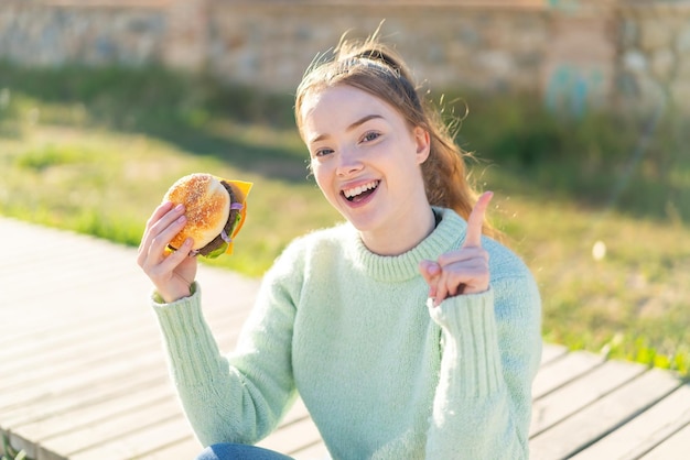 Jeune jolie fille tenant un hamburger à l'extérieur pointant vers le haut une bonne idée