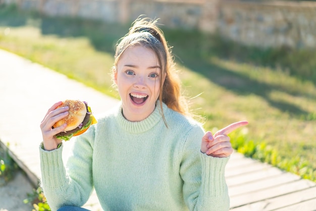 Jeune jolie fille tenant un hamburger à l'extérieur surpris et pointant le doigt sur le côté