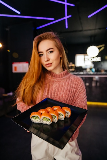 Jeune jolie fille tenant un rouleau de sushi makizushi servi sur une plaque noire sur fond de sushibar