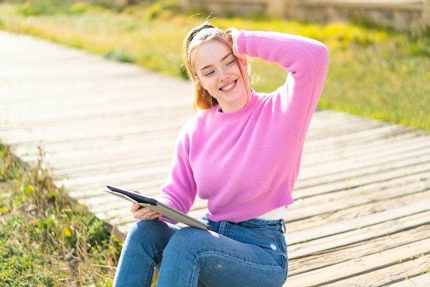 Jeune jolie fille tenant une tablette à l'extérieur ayant des doutes et avec une expression de visage confuse