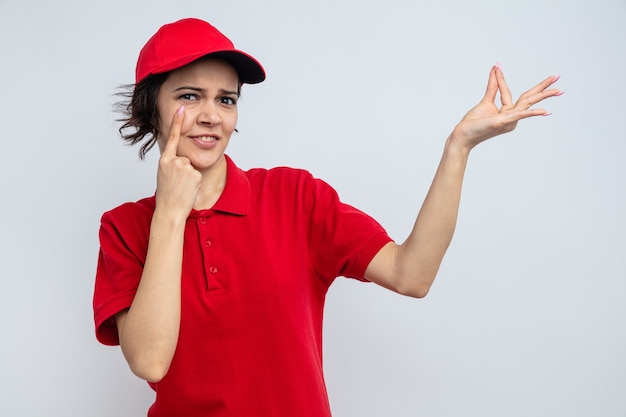 Jeune jolie livreuse impressionnée pointant du doigt son œil et faisant semblant de tenir quelque chose