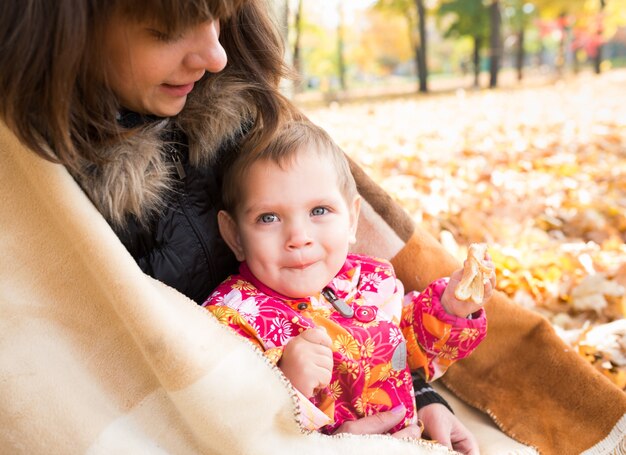 La jeune jolie maman attentionnée se ferme avec une couverture d'elle-même et de son petit enfant joyeux en marchant dans le parc d'automne. Concept de promenade de pique-nique confortable en automne