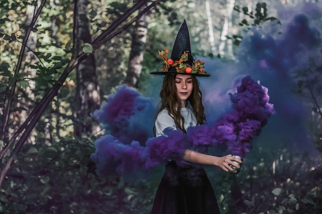 Jeune jolie sorcière au chapeau noir et avec du brouillard violet dans sa main se bouchent dans la forêt.
