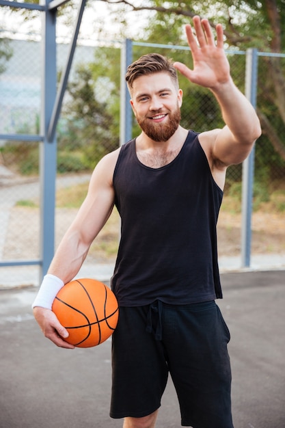 Jeune joueur de basket-ball barbu souriant saluant quelqu'un avec la main en agitant à l'extérieur