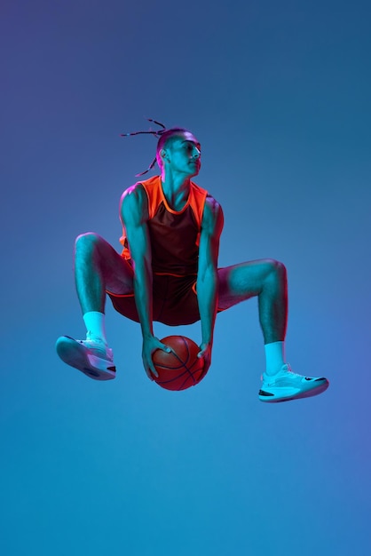 Jeune joueur de basket-ball sportif en mouvement pendant le match sautant avec le ballon contre le studio bleu