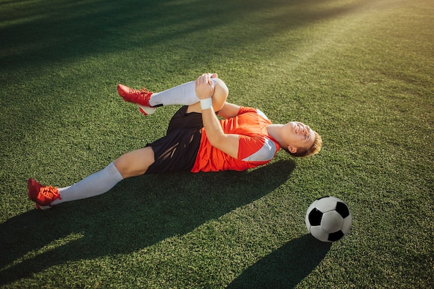 Jeune joueur de football allongé sur la pelouse et tenir la jambe. Il l'attire pour lui. Guy ressent une douleur au genou. Balle couchée à côté de lui.