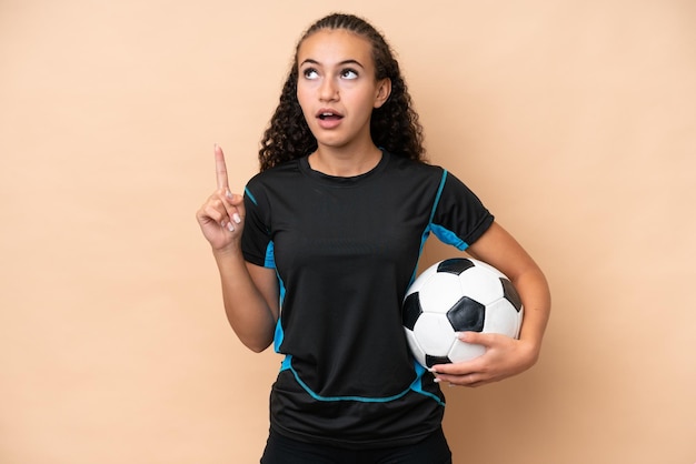 Jeune joueur de football femme isolée sur fond beige pensant une idée pointant le doigt vers le haut