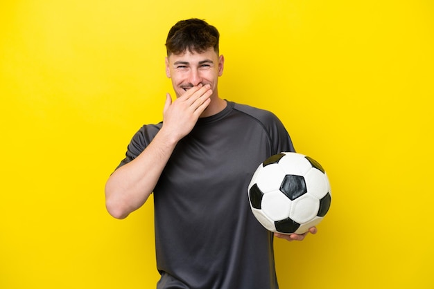 Jeune joueur de football homme isolé sur fond jaune heureux et souriant couvrant la bouche avec la main