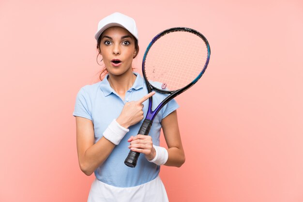 Jeune joueuse de tennis sur mur rose isolé surpris et pointant le côté