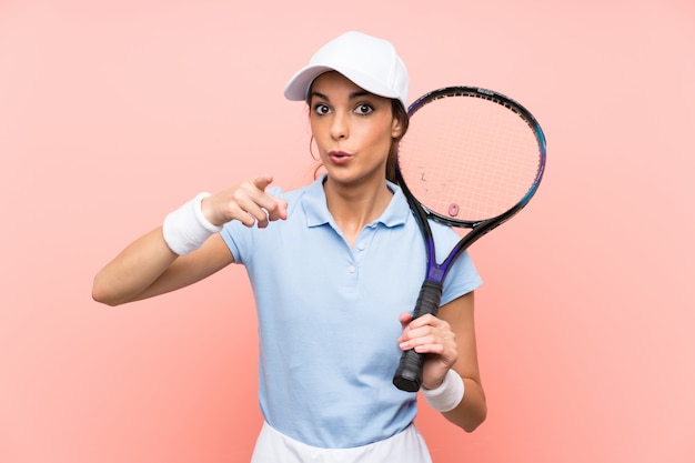 Jeune joueuse de tennis sur mur rose isolé surpris et pointant le devant