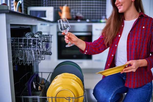 Jeune joyeuse heureuse satisfaite souriante jolie femme au foyer utilisant un lave-vaisselle moderne pour laver la vaisselle et les verres dans la cuisine de la maison blanche