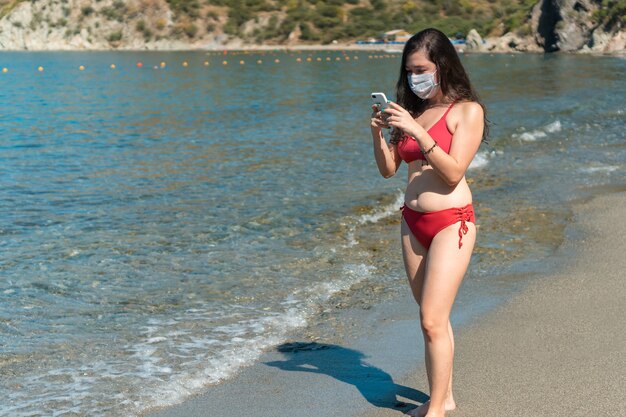 Jeune latina en masque de protection et maillot de bain vérifiant son téléphone portable