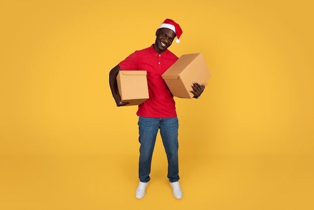 Un jeune livreur afro-américain heureux et un chapeau de Père Noël tiennent des boîtes en carton.