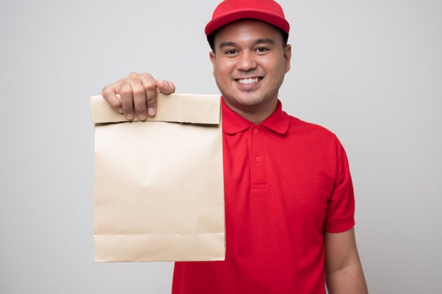 Jeune livreur asiatique souriant en uniforme rouge tenant la livraison de nourriture sac en papier