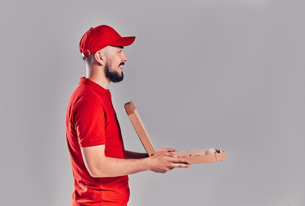 Jeune livreur barbu avec boîte à pizza isolé sur fond gris. Vue de côté.