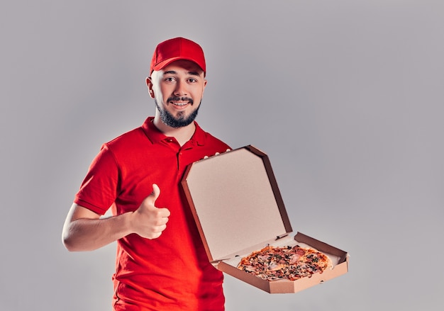 Jeune livreur barbu en uniforme rouge avec pizza montrant les pouces vers le haut geste isolé sur fond gris.