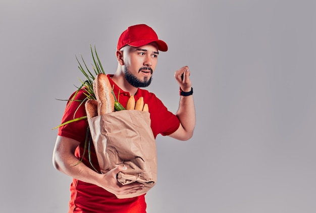 Un jeune livreur barbu en uniforme rouge tient un sac de pain et de légumes et frappe à une porte fictive isolée sur fond gris.