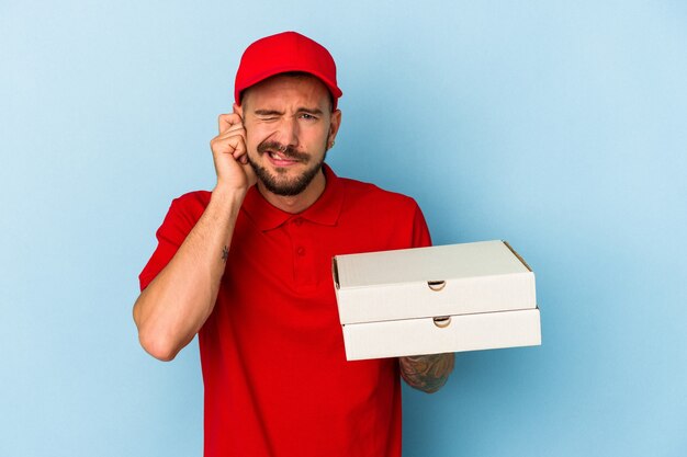 Jeune Livreur Caucasien Avec Des Tatouages Tenant Des Pizzas Isolées Sur Fond Bleu Couvrant Les Oreilles Avec Les Mains.