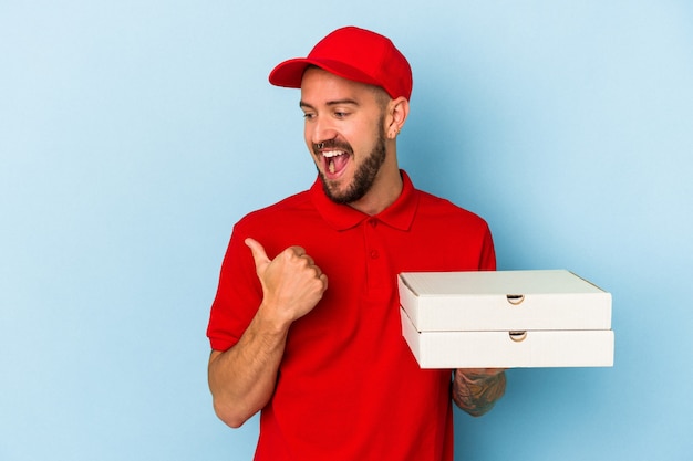 Jeune livreur caucasien avec des tatouages tenant des pizzas isolées sur des points de fond bleu avec le pouce loin, riant et insouciant.