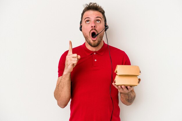 Jeune livreur caucasien tenant un hamburger isolé sur fond blanc pointant vers le haut avec la bouche ouverte.