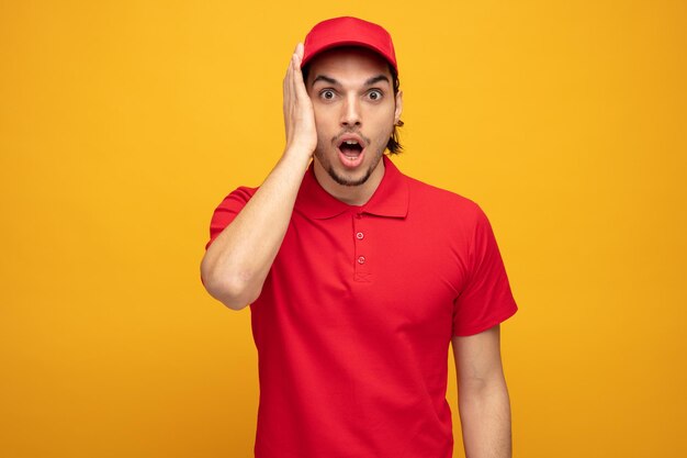 jeune livreur choqué portant l'uniforme et la casquette gardant la main sur le visage regardant la caméra isolée sur fond jaune