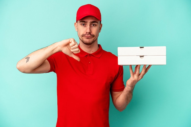 Photo jeune livreur prenant des pizzas isolées sur fond bleu montrant un geste d'aversion, les pouces vers le bas. notion de désaccord.