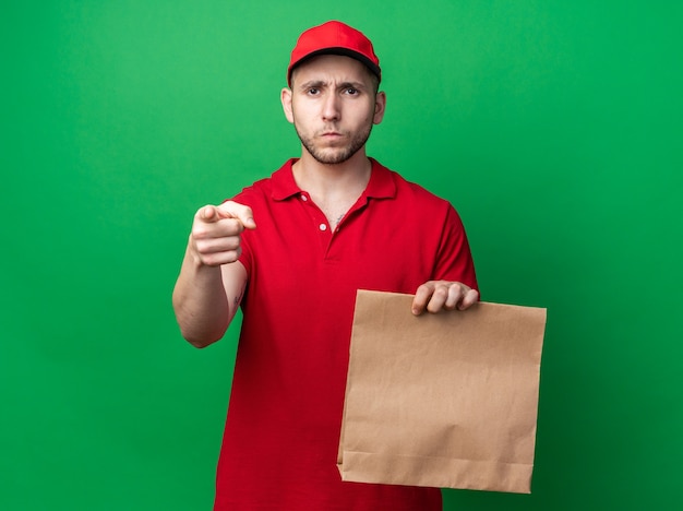 Jeune livreur strict portant un uniforme avec une casquette tenant un sac de nourriture en papier pointe à l'avant