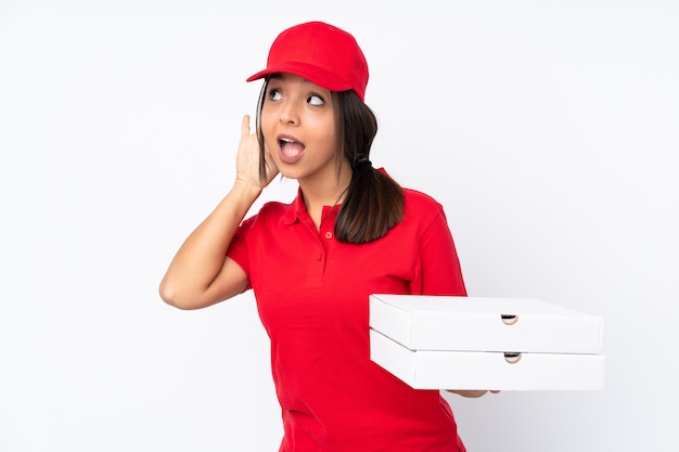 Jeune livreuse de pizza sur mur blanc isolé écouter quelque chose en mettant la main sur l'oreille