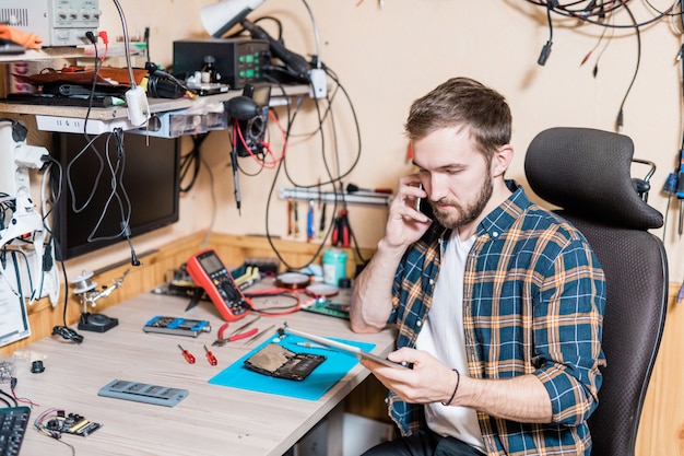 Jeune maître professionnel du service de réparation de gadgets parlant au client sur smartphone tout en regardant à travers les demandes en ligne dans le pavé tactile