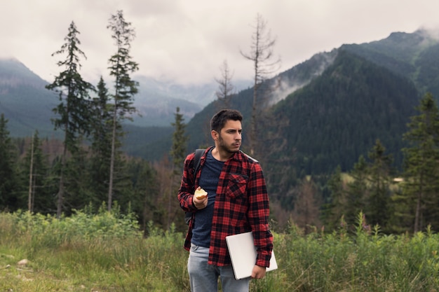 Jeune mâle brune en jeans et chemise travaille devant un ordinateur tout en étant dans la nature.