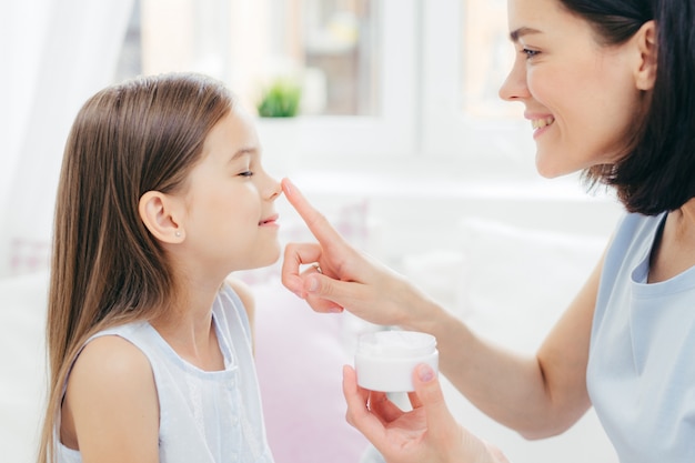 Photo jeune maman affectueuse touche le nez de sa fille, tient une bouteille de crème, apprécie la convivialité, prend soin de sa peau