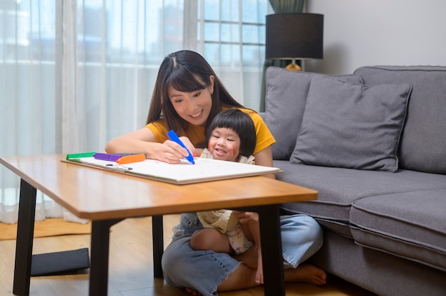 Une jeune maman aide sa fille à dessiner avec des crayons de couleur dans le salon à la maison.